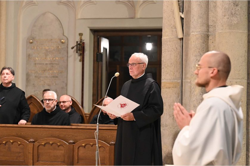 Zakonnik w czarnym habitu stoi przy smukłym mikrofonie i czyta z trzymanej w dłoniach teczki z okrągłym, czerwonym emblematem. Za nim rząd drewnianych stall z innymi zakonnikami siedzącymi i przysłuchującymi się. Wysokie kamienne filary i jasne światło kościelne dopełniają tła. Kadr podkreśla oficjalny charakter czytania.