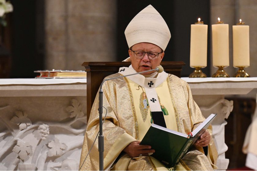 Biskup/kardynał w złotych szatach liturgicznych i białej mitrze siedzi przy ołtarzu i mówi do mikrofonu, trzymając otwartą księgę. Za nim trzy zapalone, kremowe świece ustawione w równym rzędzie; front ołtarza zdobi płytki z rzeźbionym motywem roślinnym. Wyraz twarzy poważny, skupiony; światło miękkie, ciepłe.