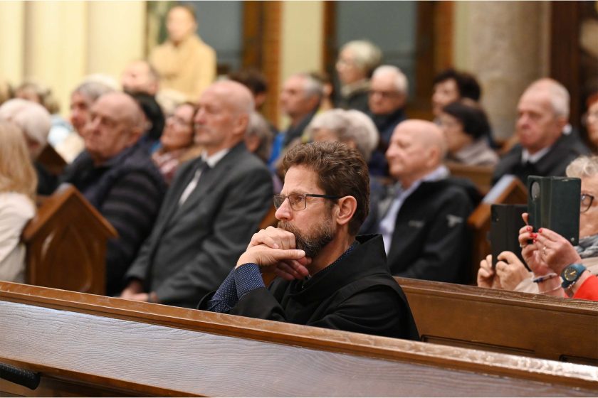 Półzbliżenie na mężczyznę w ławce kościelnej – łokieć oparty o poręcz, dłoń przy ustach, wzrok skierowany ku prezbiterium; obok i w tle wiele osób w różnym wieku, część fotografuje telefonem lub tabletem. Drewno ławek i ciepłe oświetlenie tworzą spokojny, kontemplacyjny klimat.