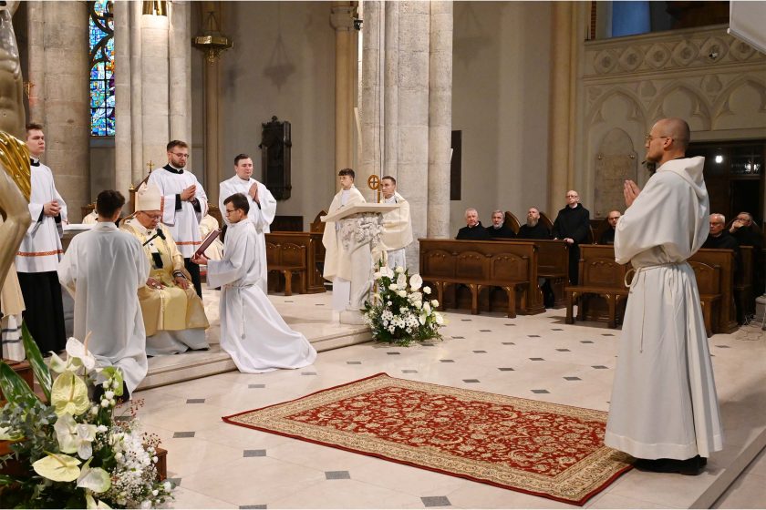 Szeroki kadr prezbiterium w trakcie obrzędu. Po lewej biskup w mitrze siedzi na katedrze; przed nim klęczy asysta liturgiczna z księgą. Po prawej stronie w pierwszym planie stoi kandydat w białym habicie, ze złożonymi dłońmi. Na posadzce, centralnie, prostokątny czerwony dywan o orientalnym wzorze; u podstawy ambony duża, biała dekoracja kwiatowa. W tle w stallach siedzą zakonnicy w czarnych habitach, obserwując przebieg ceremonii.