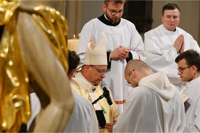 Zbliżenie na moment przysięgi/obrzędu: biskup w złotych szatach pochyla głowę, mówi do mikrofonu; przed nim, tuż przy katedrze, klęczy kandydat w białym habicie z pochyloną głową. Obok ministranci w białych albach trzymają księgi. Na lewym skraju kadru nieostry fragment pozłacanego elementu ołtarzowego. Emocja skupienia i powagi.