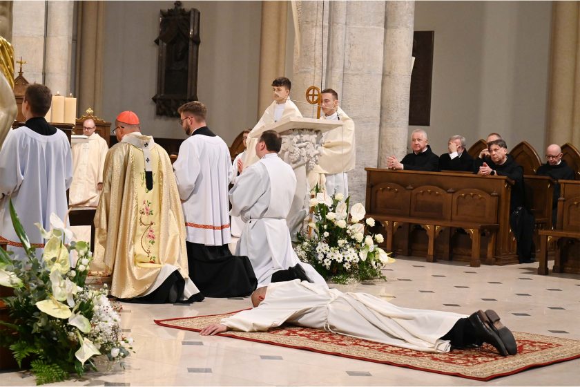 Kulminacyjny moment liturgii – prostracja. Kandydat w białym habicie leży krzyżem na czerwonym dywanie przed ołtarzem, twarzą do posadzki, ramiona wzdłuż ciała. Biskup i asysta klęczą przy ołtarzu; po prawej w stallach widać rzędy zakonników – część z dłońmi złożonymi do modlitwy, część z pochylonymi głowami. Po lewej bujna, biało-zielona dekoracja kwiatowa. Przestrzeń jasna, rytm kamiennych filarów podkreśla uroczysty charakter ceremonii.