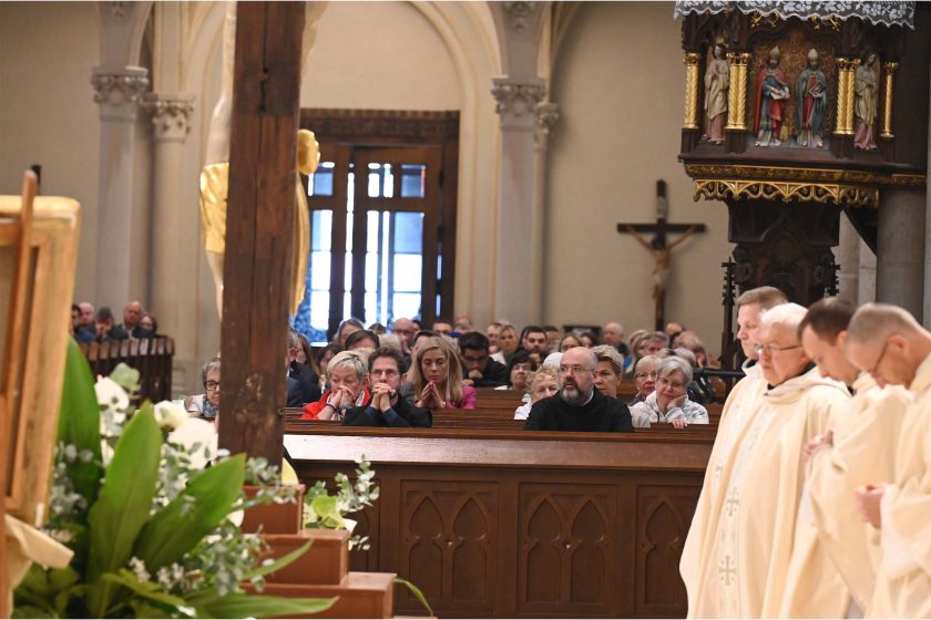 Szeroki kadr wewnątrz dużej, zabytkowej świątyni. Na pierwszym planie po lewej widać fragment masywnego, drewnianego krucyfiksu oraz biało-zieloną kompozycję kwiatową (m.in. anturia, róże, gipsówka, liście o dużych blaszkach). W głębi, po środku, ławka z wiernymi – osoby siedzą z dłońmi złożonymi do modlitwy; twarze poważne, skupione. Po prawej, przy brzegu kadru, nieostry rząd duchownych w jasnych ornatach stoi przodem do ołtarza. W tle widoczne wysokie łuki, drewniana ambona zdobiona figurkami świętych i krucyfiks na ścianie. Światło miękkie, kościelne, tworzy spokojny, uroczysty nastrój.