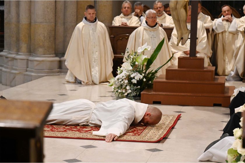 Wnętrze dużej świątyni. Na pierwszym planie zakonnik w białym habicie z kapturem leży krzyżem na czerwono-złotym, orientalnym dywanie; ręce ma wyciągnięte na boki, twarzą zwrócony do posadzki z jasnego kamienia z ciemnymi wstawkami. Za nim stoi wysoki krucyfiks na kilkustopniowym postumencie oraz duża, biała kompozycja kwiatowa (m.in. róże i anturium) z szerokimi zielonymi liśćmi. W tle w ławach siedzą i stoją kapłani w kremowych ornatach, a całość oświetla miękkie światło kościelne.