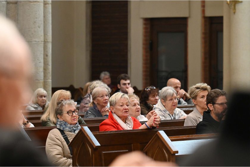 Szereg kościelnych ławek wypełnionych głównie starszymi kobietami; na środku wyróżnia się pani w czerwonym płaszczu z dłonią splecioną w geście modlitwy. Wokół osoby w okularach, w szalikach i kurtkach; wszyscy skierowani twarzami ku prezbiterium. W tle jasne filary i drewniane drzwi zakrystii; pierwszy plan częściowo rozmyty.