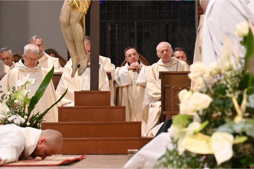 Na dole kadru widoczna głowa leżącego krzyżem zakonnika w białym habicie na czerwonym dywanie. Dominantą jest drewniany krzyż z widocznymi nogami ukrzyżowanego; po lewej bujna, biała dekoracja kwiatowa. Za postumentem siedzą kapłani w jasnych ornatach, kilku z rękami złożonymi do modlitwy; tło stanowi ciemna balustrada i arkady kościoła.