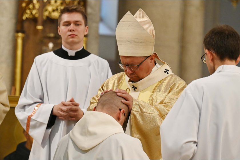W centrum biskup w złotym ornacie i mitrze pochyla głowę i oburącz kładzie dłonie na głowie klęczącego kandydata w białym habicie. Wokół stoją usługujący w białych albach, jeden trzyma księgę. Oświetlenie ciepłe, skupione; gest i wyraz twarzy podkreślają powagę obrzędu.