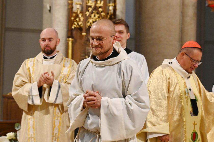 Młody zakonnik z ogoloną głową i w okularach, w jasnym habicie z kapturem, idzie przez prezbiterium z rękami złożonymi na wysokości piersi i lekkim uśmiechem. Za nim duchowny w złotych szatach liturgicznych z czerwonym zucchetto oraz inni celebransi. W tle masywne filary i elementy ołtarza.