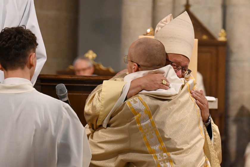 Zbliżenie: duchowny w mitrze i złotym ornacie obejmuje nowo wyświęconego (w jasnym habicie) w serdecznym uścisku. Widać pierścień na dłoni celebransa; po lewej statyw z mikrofonem. Ujęcie oddaje emocję i wspólnotowy wymiar wydarzenia.