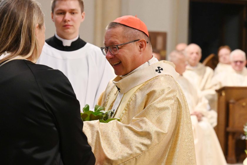 Duchowny w złotym ornacie i czerwonym zucchetto uśmiecha się szeroko, odbierając od kobiety w czerni zielony bukiet w materiale dekoracyjnym. Za nimi stoi ministrant w białej albie; w tle rozmyci kapłani i elementy prezbiterium. Światło podkreśla połysk haftów na szacie.