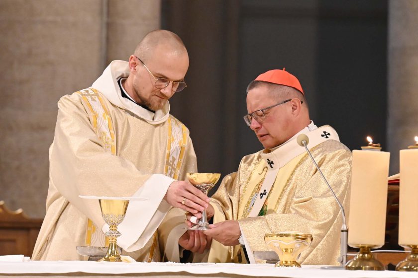 Zbliżenie na ołtarz: duchowny w dalmatyce/ornacie o złotej fakturze oraz koncelebrans w złotych szatach wspólnie trzymają i przekazują złoty kielich nad białym obrusem. W kadrze widać także patenę, drugi kielich i trzy zapalone świece na wysokich lichtarzach. Twarze skupione, gest precyzyjny.