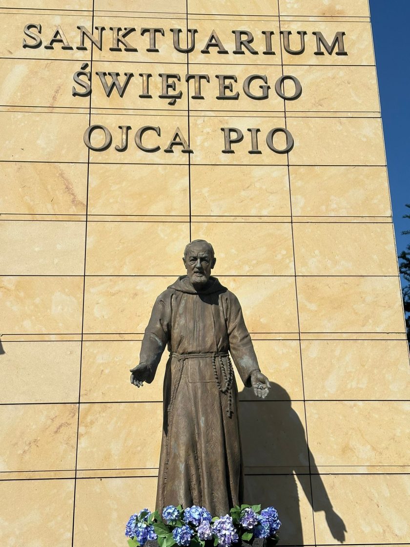 Posąg św. Ojca Pio ustawiony przed wejściem do sanktuarium, u stóp pomnika umieszczono donicę z niebieskimi hortensjami