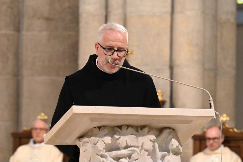 Duchowny w czarnym habicie i okularach czyta przy ambonie z jasnego kamienia zdobionej liśćmi; długi mikrofon na giętkim ramieniu skierowany do ust. Za nim, poza ostrością, widać innych kapłanów w stallach oraz masywne filary świątyni. Minę ma skupioną, wzrok opuszczony na tekst.