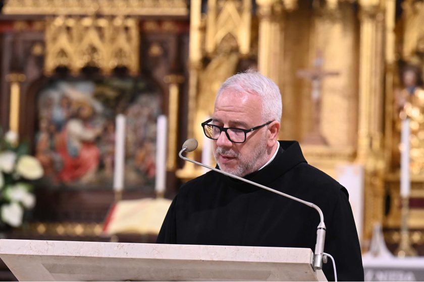 Mówca w czarnym habicie przemawia przy innej perspektywie ambony; za nim bogato złocony ołtarz z krucyfiksem i dwiema świecami, a także barwna nastawa z postaciami. Kadr półzbliżeniowy, mikrofon łukiem wychodzi z lewej, światło podkreśla siwiznę i fakturę kamienia.