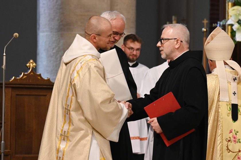 Uroczysty uścisk dłoni między nowo wyświęconym (szaty w odcieniu złamanej bieli ze złotym ornatowym pasem) a zakonnikiem w czarnym habicie trzymającym czerwony skoroszyt. Obok stoją inni duchowni; w tle widoczny fragment mitry i bogato haftowany ornat biskupa odwróconego tyłem. Nastroje pogodne, gratulacyjne.
