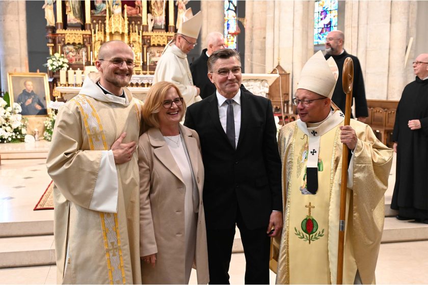 Pamiątkowe zdjęcie grupowe przed ołtarzem: po lewej nowo wyświęcony w jasnych, złocistych szatach, obok uśmiechnięta kobieta w beżowym płaszczu i mężczyzna w czarnym garniturze; po prawej biskup w mitrze z pastorałem, uśmiechnięty. W tle monumentalna, złocona nastawa ołtarzowa oraz obraz świętego na sztaludze, a po bokach krzątający się duchowni.