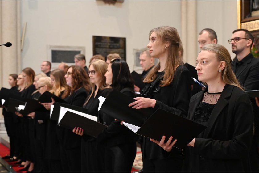 Rząd chórzystów ustawionych ukośnie względem obiektywu; wszyscy w czarnych stylizacjach (marynarki, bluzki, koszule) i z nutami. Dwie kobiety na pierwszym planie w półprofilu, wyraźna faktura materiałów i detale (kolczyki, falujące włosy). Głębia pola średnia — twarze z przodu ostre, dalsze sylwetki łagodnie rozmyte.