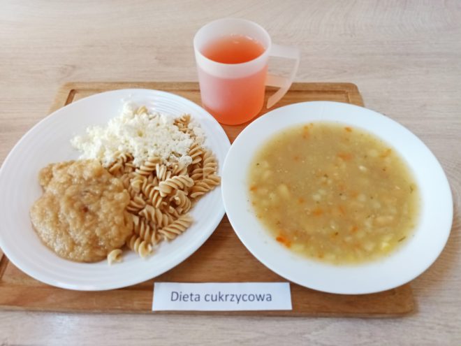 Na tacy podano dwudaniowy obiad. W głębokim talerzu znajduje się zupa jarzynowa z kawałkami warzyw. Na drugim talerzu ułożono porcję pełnoziarnistego makaronu świderki, białą surówkę z kapusty oraz kawałek pieczonego mięsa w sosie. Do posiłku podano kubek z kompotem owocowym bez dodatku cukru. Etykieta na tacy informuje „Dieta cukrzycowa”