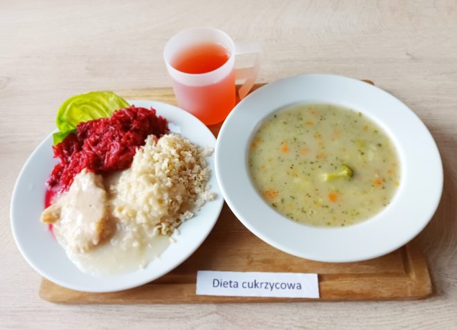 Na drewnianej tacy ustawiony zestaw obiadowy: biały talerz z kaszą, duszonym mięsem w jasnym sosie, buraczkami i liściem sałaty. Obok miska z zupą jarzynową z kawałkami brokuła oraz kubek z czerwonym napojem. Przed tacą kartka z napisem „Dieta cukrzycowa”