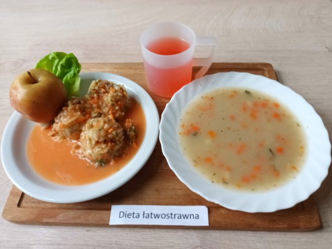 Na tacy widoczny jest zestaw obiadowy w diecie łatwostrawnej: talerz z zupą jarzynową na białym talerzu, porcja gołąbków w sosie pomidorowym z dodatkiem liścia sałaty i jabłka, obok przezroczysty kubek z różowym kompotem