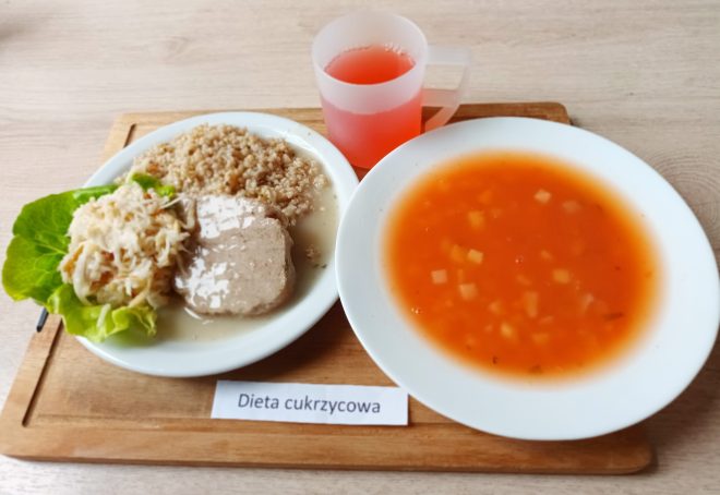 Na drewnianej tacy znajduje się posiłek oznaczony jako dieta cukrzycowa. Po lewej stronie talerza widoczny jest plaster gotowanego mięsa w jasnym sosie, obok porcja kaszy i surówka z kapusty na liściu sałaty. Po prawej stronie tacy stoi głęboki talerz z zupą pomidorową z kawałkami warzyw. Obok ustawiono przezroczysty kubek z różowym napojem