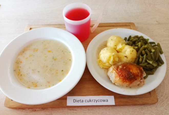 Na drewnianej tacy znajduje się zestaw obiadowy dla osób na diecie cukrzycowej. Po lewej stronie w białej misce podana jest jasna zupa jarzynowa z dodatkiem marchewki i przypraw. Po prawej stronie, na talerzu, widać porcję gotowanych ziemniaków z natką pietruszki, ugotowaną na parze fasolkę szparagową oraz kawałek pieczonego kurczaka z chrupiącą skórką. Do zestawu dołączono przezroczysty kubek z czerwonym kompotem owocowym