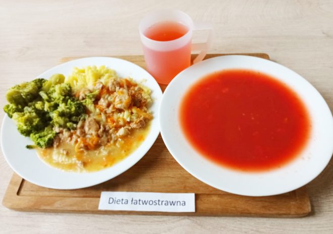 Na tacy ustawiono zestaw obiadowy w ramach diety łatwostrawnej. Na dużym białym talerzu znajduje się porcja zupy pomidorowej o jednolitej, czerwonej konsystencji. Obok, na drugim talerzu, podano gotowane ziemniaki z sosem i mięsem duszonym z warzywami, a także porcję brokułów. Do posiłku dołączono przezroczysty kubek z czerwonym napojem owocowym