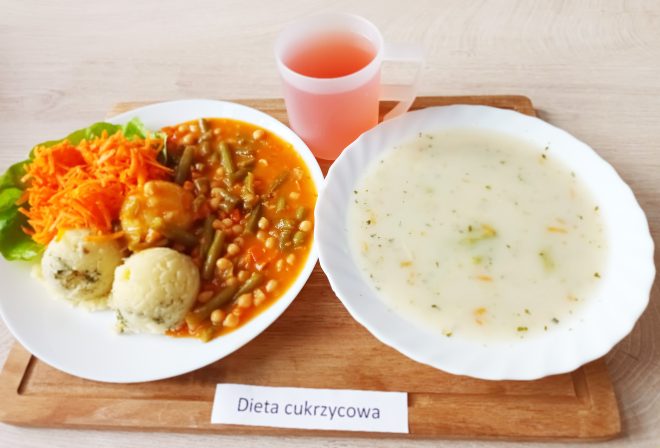 Na drewnianej tacy ustawiono zestaw obiadowy opisany jako „Dieta cukrzycowa”. Na talerzu znajdują się dwie kulki ziemniaków z koperkiem, surówka z marchewki na liściu sałaty oraz gulasz warzywny z fasolką szparagową i groszkiem w sosie pomidorowym. Obok podano biały, głęboki talerz z zupą jarzynową na bazie białego wywaru. Do zestawu dołączono kubek z kompotem
