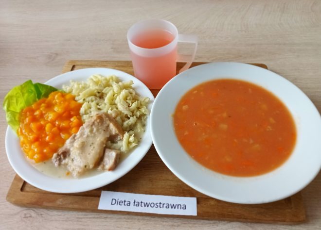 Na tacy znajduje się zupa jarzynowa w białym talerzu, drugi talerz z gotowanym mięsem w sosie, makaronem świderki i gotowaną marchewką. Do posiłku podany różowy kompot w plastikowym kubku. Kartka z podpisem „Dieta łatwostrawna”