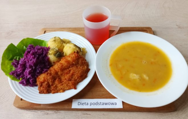 Na drewnianej tacy znajduje się posiłek oznaczony jako „Dieta podstawowa”. Na jednym talerzu podane są ziemniaki z koperkiem, surówka z czerwonej kapusty, liść sałaty oraz panierowany kotlet schabowy. Obok znajduje się kubek z napojem i miska z żółtą zupą jarzynową