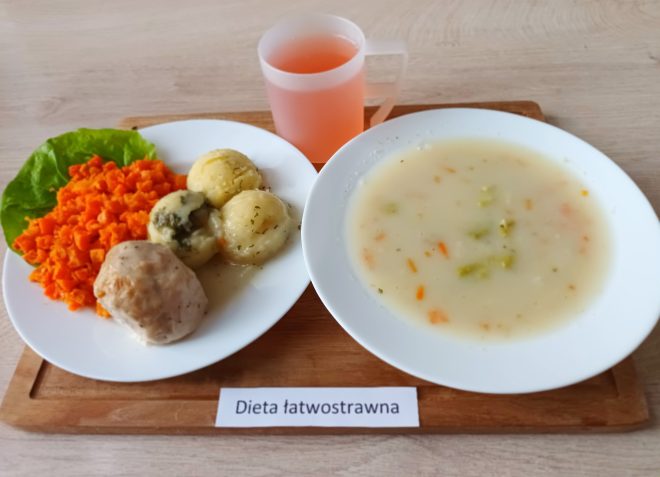 Taca z zestawem obiadowym w diecie łatwostrawnej: talerz z gotowaną marchewką, puree ziemniaczanym w formie kulek z sosem, pulpetem drobiowym oraz liściem sałaty; obok miseczka z zupą jarzynową na białej bazie i kubek z różowym napojem