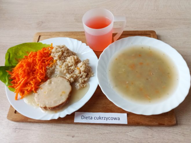 Obiad w diecie cukrzycowej: talerz z gotowaną kaszą, plasterkiem mięsa w sosie, surówką z tartej marchewki i listkiem sałaty. Obok miska z lekką zupą jarzynową oraz kubek z kompotem owocowym