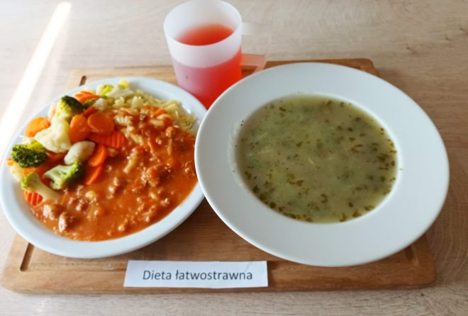 Na zdjęciu widoczny jest zestaw obiadowy oznaczony etykietą „Dieta łatwostrawna”. Na tacy ustawiono biały talerz z zupą jarzynową o jasnym wywarze, obok znajduje się drugi talerz z makaronem polanym sosem mięsnym oraz gotowanymi warzywami, takimi jak marchew, brokuł i kalafior. Do posiłku podano również napój w plastikowym kubku o czerwonym kolorze
