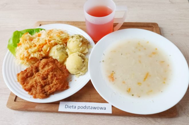 Na dużym talerzu podano panierowany kotlet schabowy, porcję ziemniaków z koperkiem oraz surówkę z kapusty i marchwi. Obok znajduje się talerz z zupą jarzynową oraz kubek z czerwonym napojem. Całość ustawiona na tacy z karteczką „Dieta podstawowa”