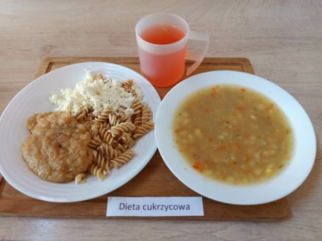 Na tacy umieszczono obiad składający się z talerza z makaronem pełnoziarnistym, startym białym serem i porcją duszonego mięsa w sosie. Obok stoi miska z zupą jarzynową na bazie ziemniaków i marchwi. Do posiłku dołączono kubek z napojem owocowym. Przed tacą znajduje się kartka z napisem „Dieta cukrzycowa”