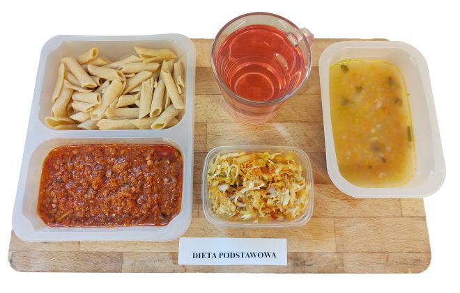 Na tacy znajduje się zestaw obiadowy w pojemnikach. W jednym z nich podano makaron penne, w drugim sos pomidorowy z mięsem mielonym, a w kolejnym surówkę z kapusty z dodatkiem marchewki. Obok znajduje się zupa jarzynowa w plastikowym pojemniku oraz szklanka kompotu. Na tacy widoczna jest etykieta „Dieta podstawowa”