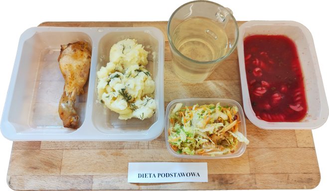 Zestaw obiadowy w ramach diety podstawowej podany na tacy. W plastikowych pojemnikach znajduje się pieczona pałka z kurczaka, porcja ziemniaków puree posypanych koperkiem oraz barszcz czerwony z makaronem. Obok podano surówkę z kapusty pekińskiej z marchewką i dodatkami. Do posiłku dołączono szklankę jasnego napoju