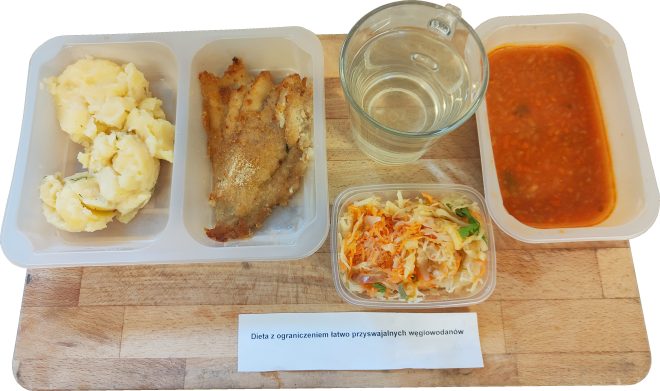 Taca z podziałkami: porcja gotowanych ziemniaków z koperkiem, panierowany filet rybny, surówka z kapusty i marchewki. Obok szklanka wody oraz zupa jarzynowa w rosole pomidorowym. Na kartce napis „Dieta z ograniczeniem łatwo przyswajalnych węglowodanów”