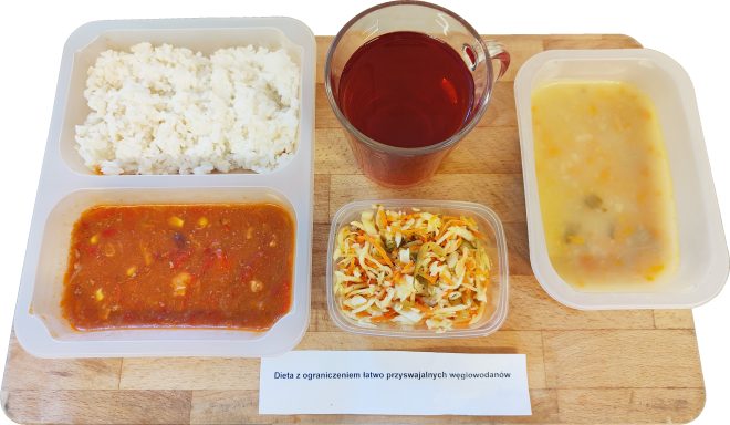 Na drewnianej tacy znajduje się zestaw obiadowy opisany jako „Dieta z ograniczeniem łatwo przyswajalnych węglowodanów”. W dużym pojemniku podano porcję ryżu oraz gulasz warzywno-mięsny w sosie pomidorowym z dodatkiem papryki i kukurydzy. Obok ustawiono mniejszy pojemnik z surówką z kapusty i marchwi. Z boku znajduje się pojemnik z zupą jarzynową o jasnej, lekko zagęszczonej konsystencji. Do posiłku podano szklankę czerwonego kompotu