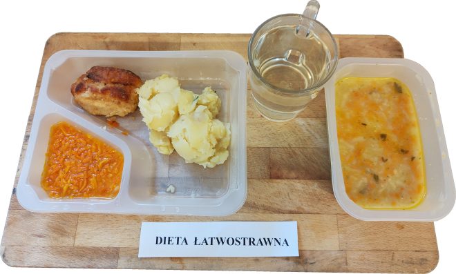 Porcjowany pojemnik z gotowaną marchewką, kotletem mielonym i ziemniakami. Obok szklanka z wodą oraz zupa jarzynowa w drugim pojemniku. Na tacy kartka z napisem „Dieta łatwostrawna”