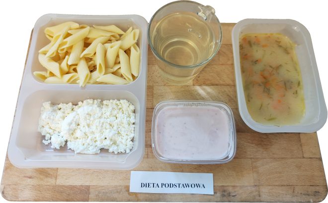 Na drewnianej desce ustawiono zestaw obiadowy w pojemnikach. Widzimy porcję gotowanego makaronu penne, twaróg, kubek z jasnym napojem (prawdopodobnie herbata lub kompot), miseczkę zupy jarzynowej z koperkiem oraz deser w postaci jogurtu owocowego. Na kartce znajduje się napis „DIETA PODSTAWOWA”