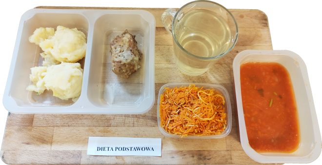 Taca z obiadem w diecie podstawowej: w plastikowym pojemniku dwie porcje ziemniaków puree oraz kawałek pieczeni mielonej. Obok porcja surówki z marchewki, miseczka z zupą pomidorową oraz szklanka z jasnym napojem