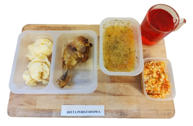 Zestaw obiadowy w plastikowych pojemnikach na desce drewnianej, oznaczony etykietą „Dieta podstawowa”. W skład posiłku wchodzą: porcja puree ziemniaczanego, pieczone udko kurczaka, zupa jarzynowa z makaronem, surówka z białej kapusty i marchewki, szklanka czerwonego napoju owocowego