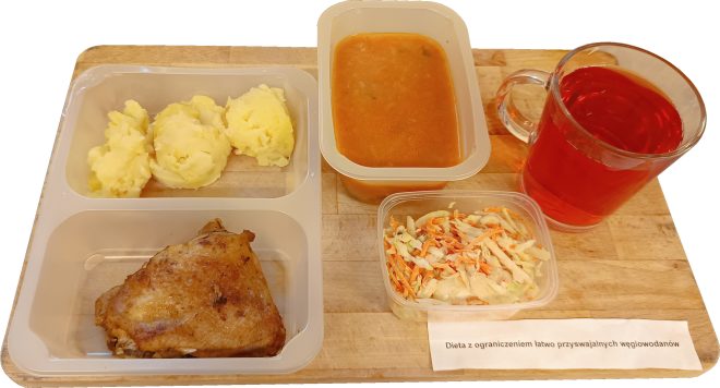 Na drewnianej tacy znajduje się zestaw obiadowy opisany jako „Dieta z ograniczeniem łatwo przyswajalnych węglowodanów”. Widać porcję puree ziemniaczanego, pieczone udko kurczaka, surówkę z kapusty i marchewki w plastikowym pojemniczku, zupę pomidorową w pojemniku oraz szklankę czerwonego napoju.