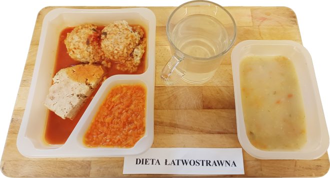 Na tacy umieszczono posiłek oznaczony jako „Dieta łatwostrawna”. W pojemniku znajduje się kawałek klopsa w sosie pomidorowym z kaszą, dwa pulpety ryżowe w sosie pomidorowym oraz duszona marchewka. Do tego podano lekką zupę jarzynową w osobnym pojemniku oraz szklankę wody.