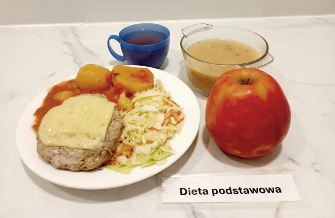 Zestaw obiadowy w diecie podstawowej: kotlet mielony zapiekany z serem, gotowane ziemniaki z sosem pomidorowym, surówka z kapusty, zupa jarzynowa, jabłko oraz herbata w niebieskim kubku
