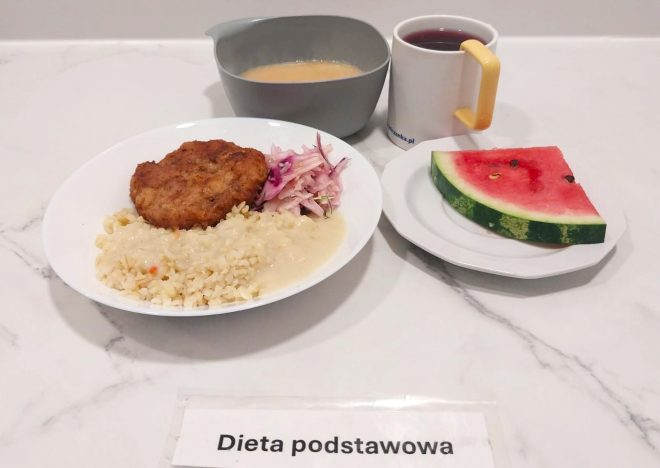 Porcja ryżu polanego jasnym sosem, kotlet mielony panierowany i surówka z czerwonej kapusty na białym talerzu. Do zestawu miseczka z zupą kremem, kubek z herbatą oraz kawałek arbuza na małym talerzyku. Przed talerzem widoczna kartka z napisem „Dieta podstawowa”