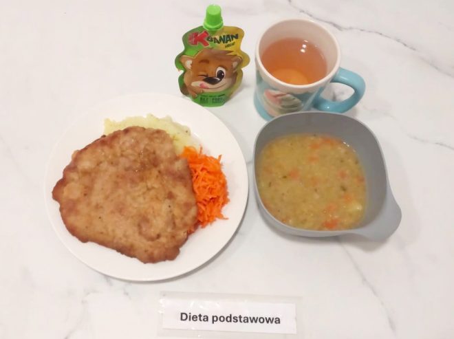 Na stole ustawiono obiad w ramach diety podstawowej. Na białym talerzu znajduje się panierowany kotlet schabowy, porcja puree ziemniaczanego oraz surówka z marchwi. Obok stoi miseczka z zupą jarzynową na bazie kaszy, z widocznymi kawałkami marchewki i ziemniaka. Do posiłku dołączono kubek z ciepłym napojem oraz mus owocowy w tubce z zielonym korkiem