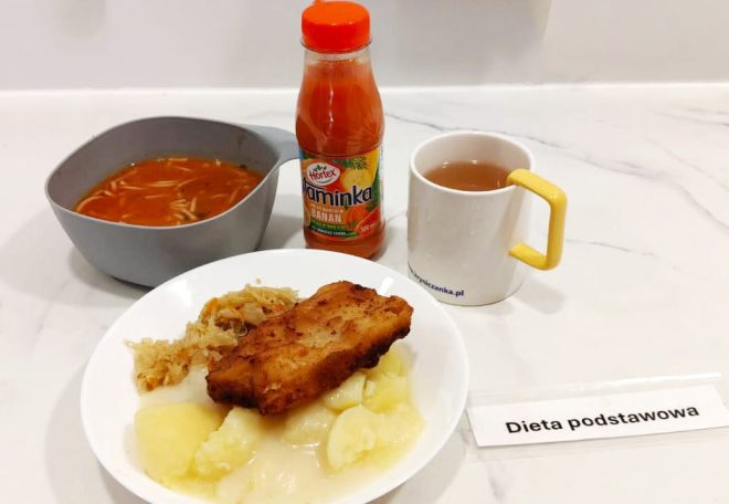 Na talerzu puree ziemniaczane z kapustą zasmażaną i panierowaną rybą. Obok miseczka z zupą pomidorową z makaronem, kubek z herbatą i butelka soku „Vitamina Hortex”. Na stole kartka z napisem „Dieta podstawowa”