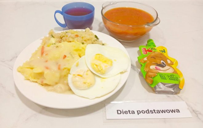 Na białym talerzu znajduje się porcja ziemniaków puree polanych sosem jarzynowym, dwie połówki jajka na twardo oraz surówka z kapusty. Obok ustawiono szklaną miskę z zupą pomidorową oraz niebieski kubek z herbatą. Do zestawu dołączono owocowy mus w saszetce z rysunkiem misia. Całość opisana jest jako „Dieta podstawowa”